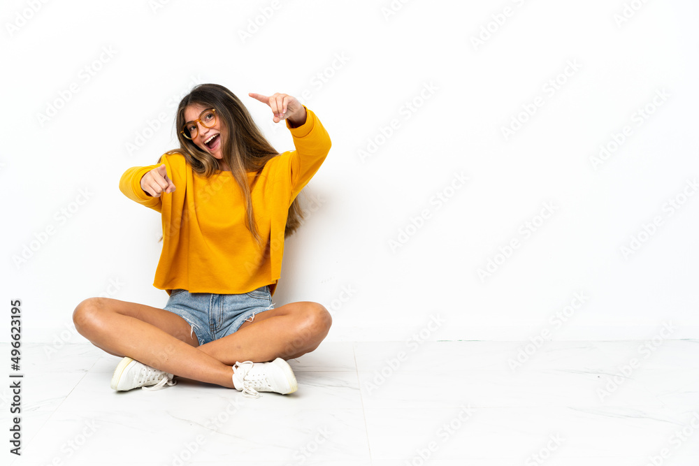 Young woman sitting on the floor isolated on white background points finger at you while smiling Young woman sitting on the floor isolated on white background points finger at you while smiling