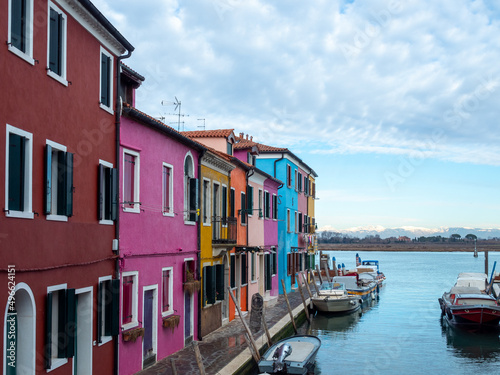 A row of brightly colored houses face the canal on the beautiful Venetian island of Burano, the snow-capped Dolomite mountains are in the background - Burano, Venice, Italy