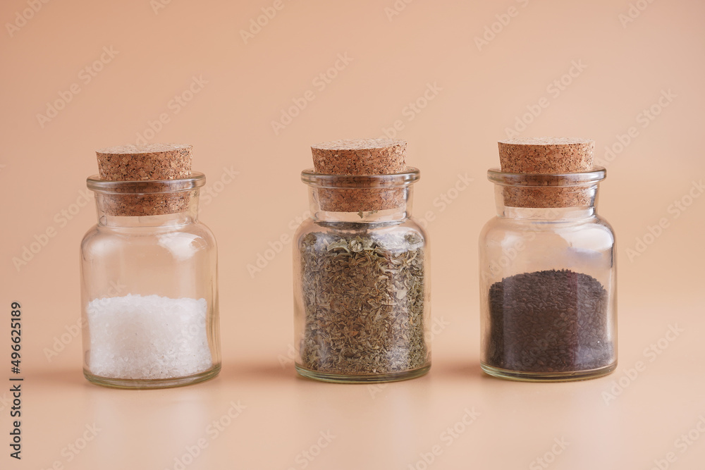 Three glass jars with cork lids containing spices: coarse sea salt, oregano, black sesame seeds on yellow seamless surface Three glass jars with cork lids containing spices: coarse sea salt, oregano, black sesame seeds on yellow seamless surface