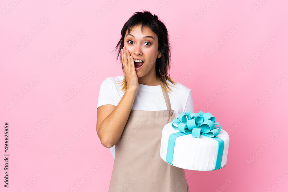 Uruguayan Pastry chef holding a big cake over isolated pink background with surprise facial expression Uruguayan Pastry chef holding a big cake over isolated pink background with surprise facial expression
