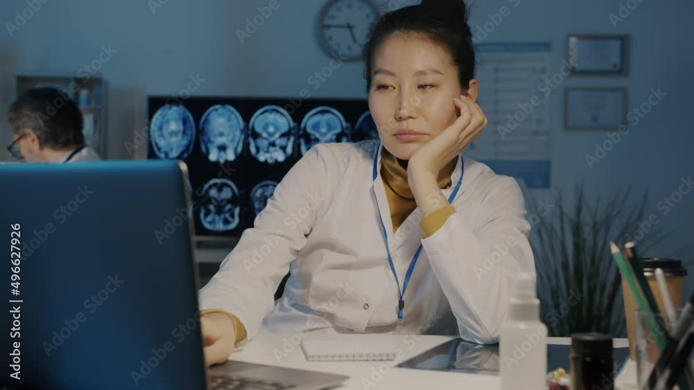 Slow motion portrait of tired Asian lady doctor in white coat using laptop and napping in dark office at night. Medicine and overwork concept. Slow motion portrait of tired Asian lady doctor in white coat using laptop and napping in dark office at night. Medicine and overwork concept.