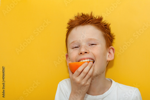 A red-haired boy appetizingly bites half of a juicy orange on a yellow background