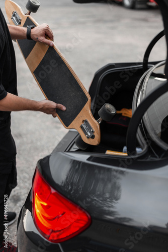 Cool man in black clothes takes a wooden longboat out of the trunk of a black car. Active recreation concept