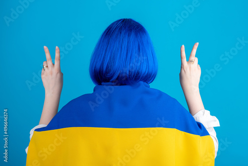 Support Ukraine concept. Woman with short hair blue wig, white blouse and Ukraine national flag on her shoulders standing with her hands up with v or victory finger signs in blue background