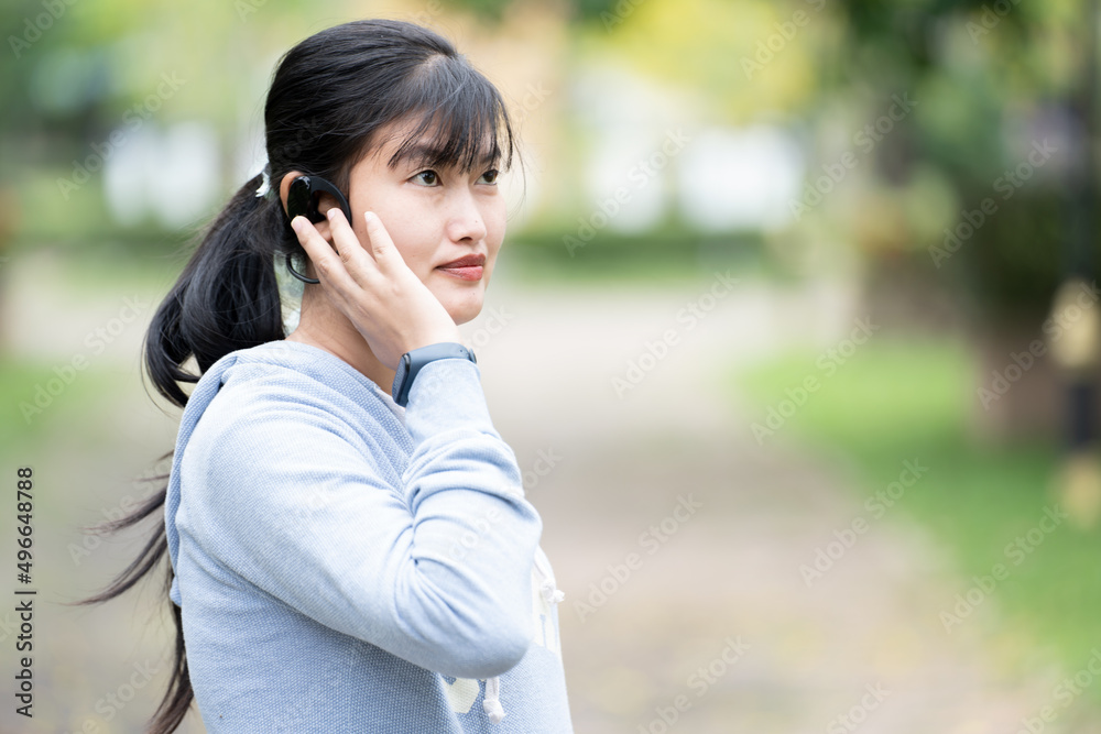 A young woman wearing a long-sleeved shirt is listening to music with Bluetooth headphones in hand with a smart watch, getting ready for a workout at the park. A young woman wearing a long-sleeved shirt is listening to music with Bluetooth headphones in hand with a smart watch, getting ready for a workout at the park.