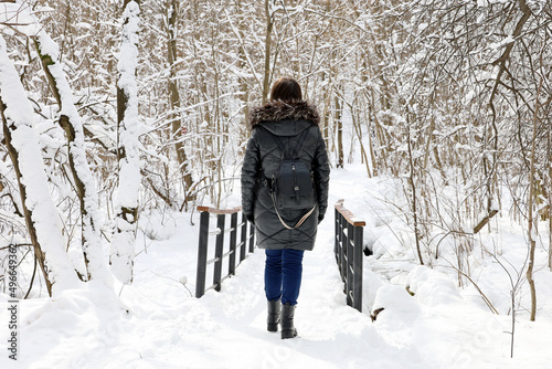 Lonely woman walking on a wooden bridge covered with snow. Winter or early spring park after snowfall, leisure on nature in cold weather