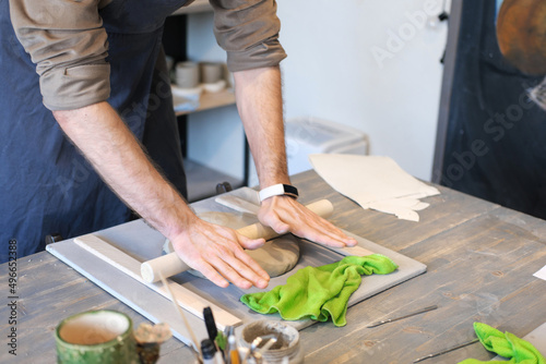 A close-up of a man potter rolls a brown clay rolling pin on a special fabric on a wooden table to make a plate