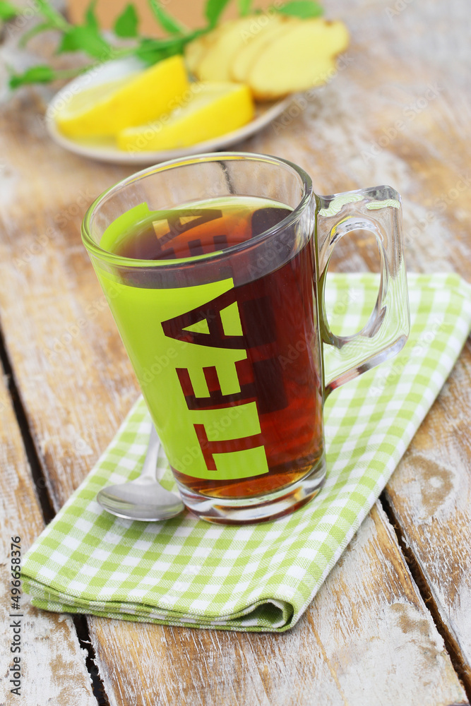 Black tea in transparent glass on checkered cloth with slices of fresh ginger, lemon and mint in the background