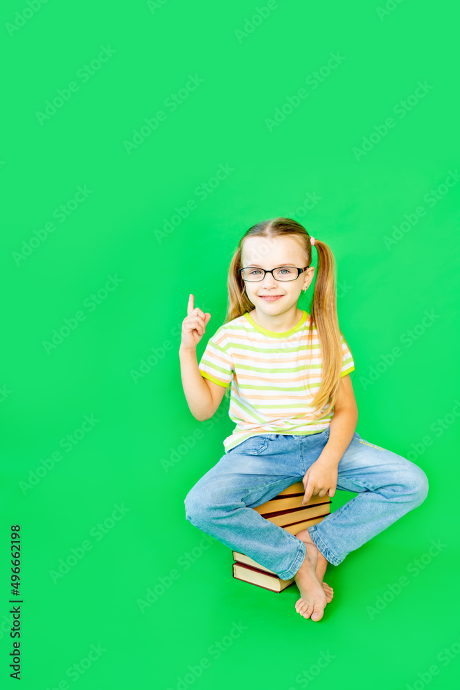 a girl child sits on a green background on books with glasses and points her finger up or to the side, space for text a girl child sits on a green background on books with glasses and points her finger up or to the side, space for text