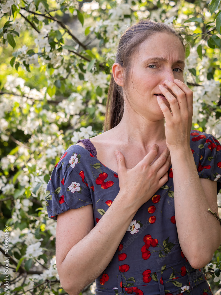 Young woman sneezes in the park against the background of a flowering tree. Allergy to pollen concept. Young woman sneezes in the park against the background of a flowering tree. Allergy to pollen concept.