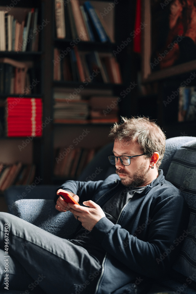 Man wearing glasses using a smartphone while sitting on a couch in the library with books in the background. College life. Man wearing glasses using a smartphone while sitting on a couch in the library with books in the background. College life.