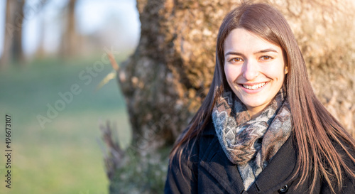 Portrait of young brunette caucasian woman. She is sitting at the foot of a tree on a bright spring day. She smiles looking at the camera.