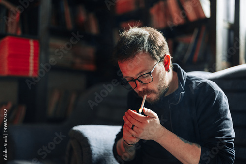 Smoking Male student wearing glasses in library with bookshelves on background.