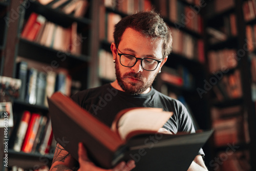 Adult student reading a book in the college library. Young positive male wearing glasses with bookshelves on background.