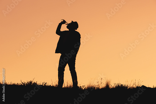 silhouette of man drinking water from bottle. man in hat stands and drinks water