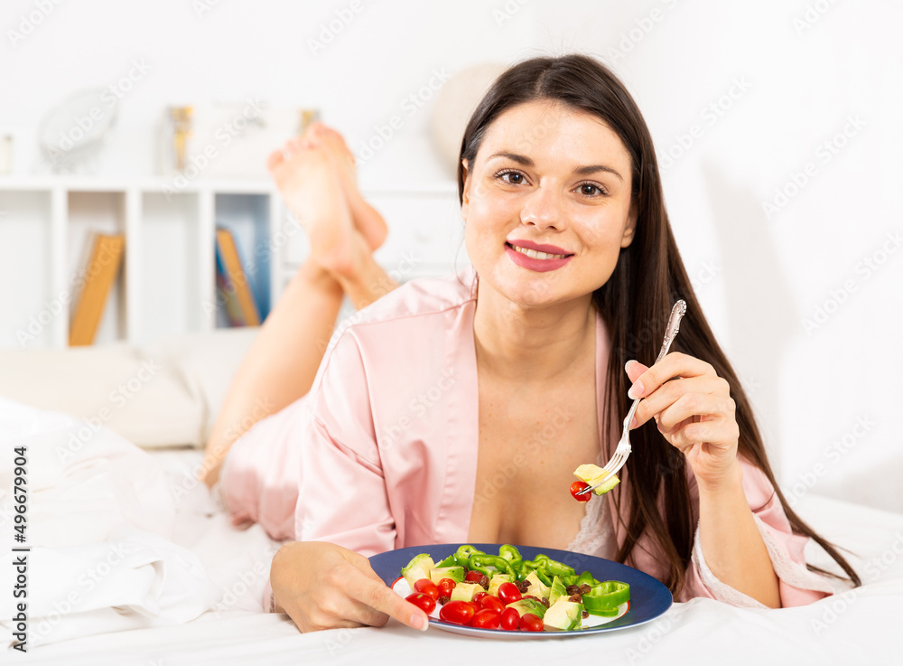 Young pretty woman in bathrobe eating vegetable salad from plato in bed at home
