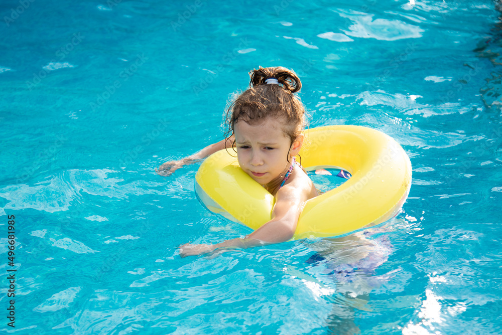 A beautiful little smiling girl in a swimsuit and in a yellow inflatable circle bathes and plays in the pool with clear blue water. Summer. Vacation. Rest.