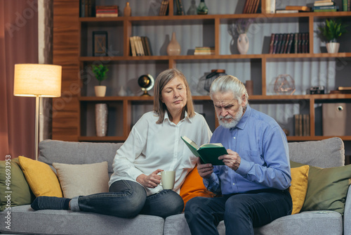 senior couple spending leisure time reading a book together sitting on cozy sofa at home. Elderly mature family Gray hair man and woman together carefree retirement lifestyle. Cozy in house