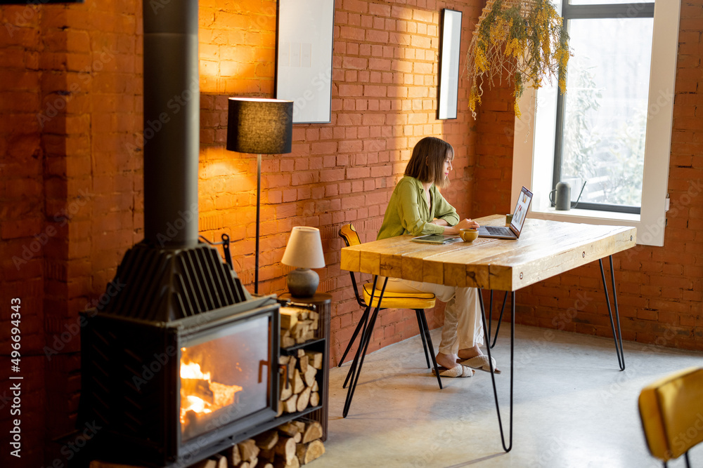 Woman works on laptop while sitting by the wooden table in cozy living room with a burning fireplace in front. Interior in loft style Woman works on laptop while sitting by the wooden table in cozy living room with a burning fireplace in front. Interior in loft style