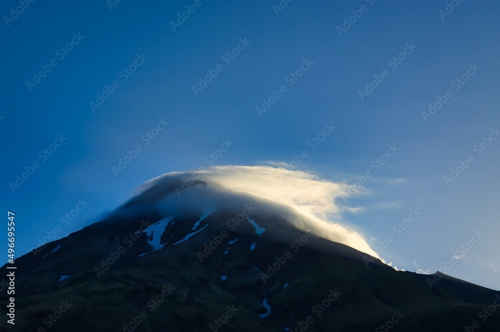 Volcano with snowfields and clouds around the summit, lit by the evening sun. Mount Taranaki (Mount Egmont), North Island, New Zealand.
