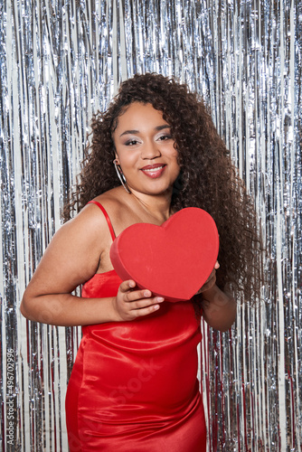 Multiracial woman in luxurious red dress holding present for the Valentines Day while posing
