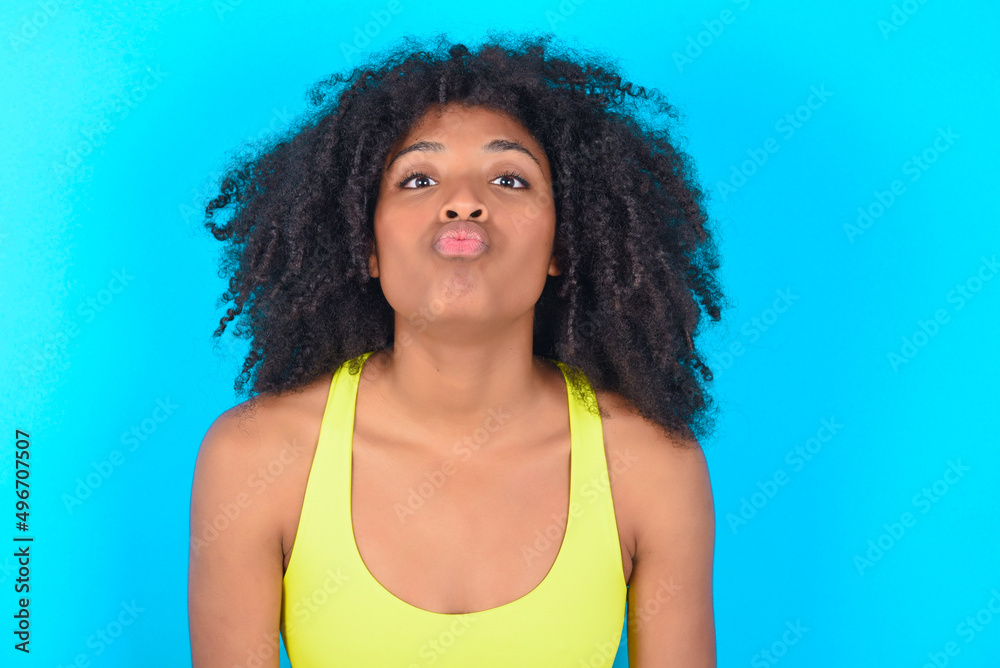 young woman with afro hairstyle in sportswear against blue background, keeps lips as going to kiss someone, has glad expression, grimace face. Standing indoors. Beauty concept. young woman with afro hairstyle in sportswear against blue background, keeps lips as going to kiss someone, has glad expression, grimace face. Standing indoors. Beauty concept.