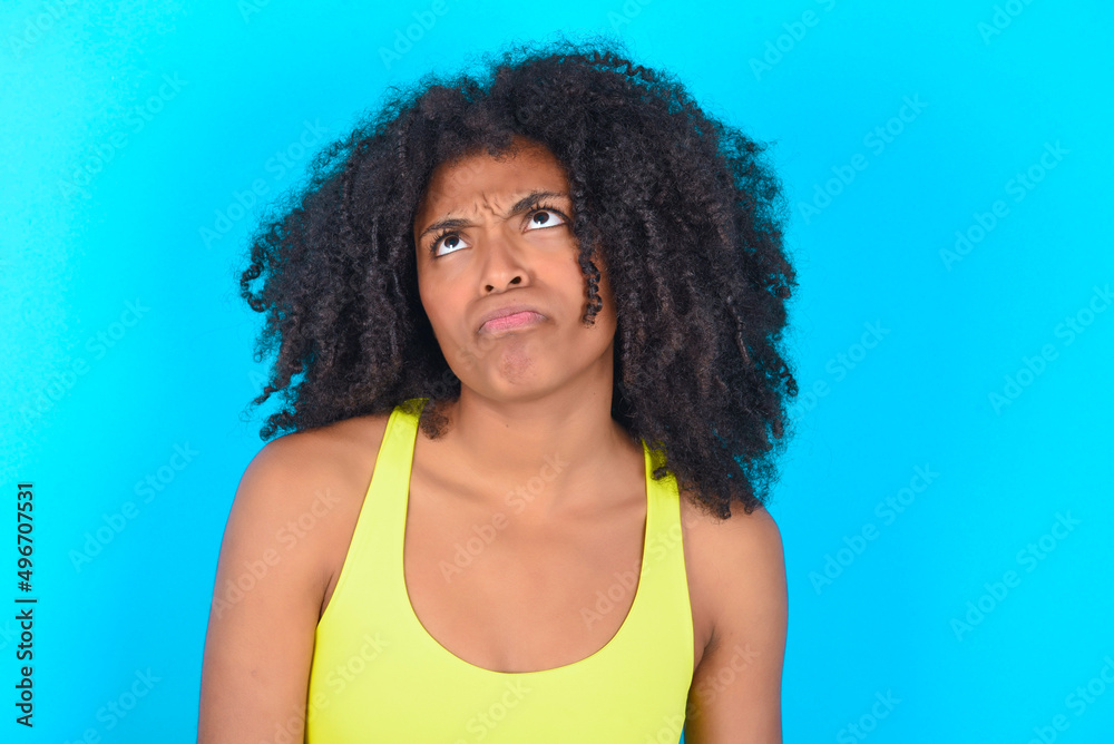young woman with afro hairstyle in sportswear against blue background has worried face looking up lips together, being upset thinking about something important, keeps hands down. young woman with afro hairstyle in sportswear against blue background has worried face looking up lips together, being upset thinking about something important, keeps hands down.