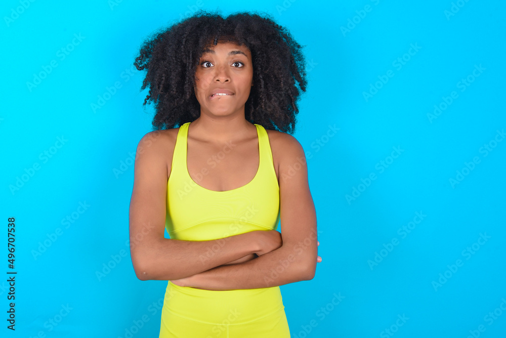 young woman with afro hairstyle in sportswear against blue background bitting his mouth and looking worried and scared crossing arms, worry and doubt. young woman with afro hairstyle in sportswear against blue background bitting his mouth and looking worried and scared crossing arms, worry and doubt.