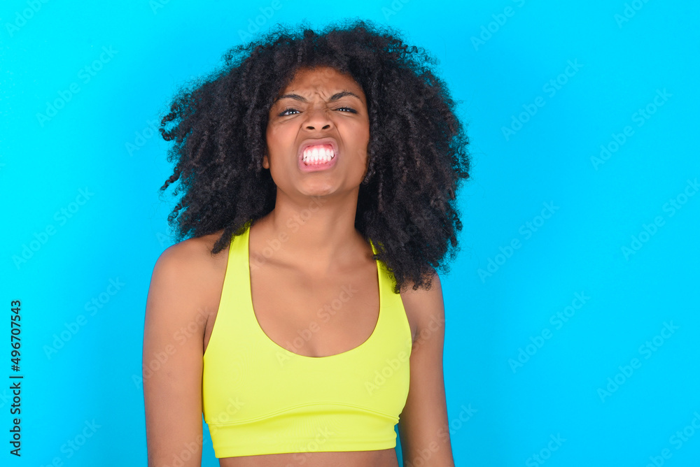 Mad crazy young woman with afro hairstyle in sportswear against blue background clenches teeth angrily, being annoyed with coming noise. Negative feeling concept.