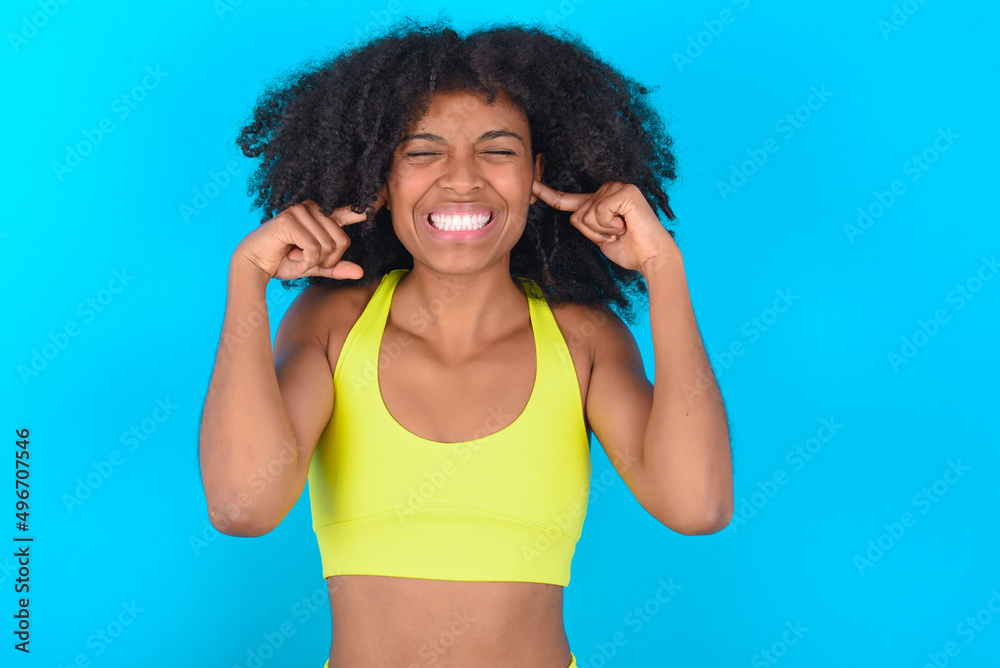 Stop making this annoying sound! Unhappy stressed out young woman with afro hairstyle in sportswear against blue background making worry face, plugging ears with fingers, irritated with loud noise.