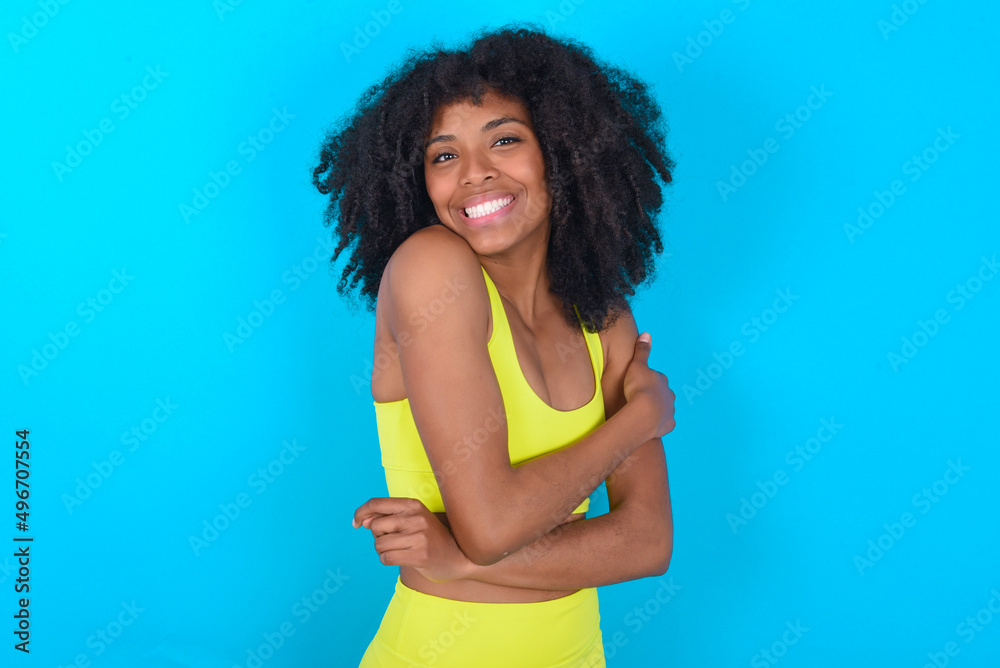People, lifestyle, youth and happiness concept. Shy pretty young woman with afro hairstyle in sportswear against blue background, feeling happy hugging herself. People, lifestyle, youth and happiness concept. Shy pretty young woman with afro hairstyle in sportswear against blue background, feeling happy hugging herself.