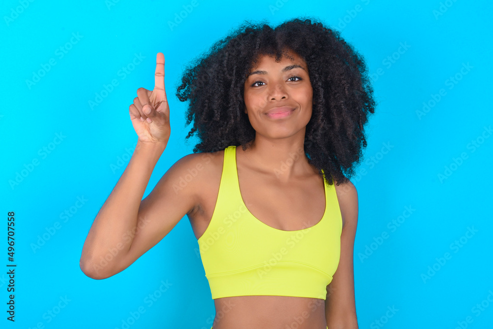 young woman with afro hairstyle in sportswear against blue background showing and pointing up with fingers number one while smiling confident and happy.