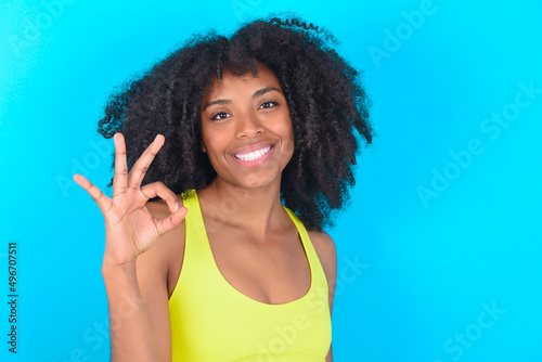 Glad attractive young woman with afro hairstyle in sportswear against blue background shows ok sign with hand as expresses approval, has cheerful expression, being optimistic.
