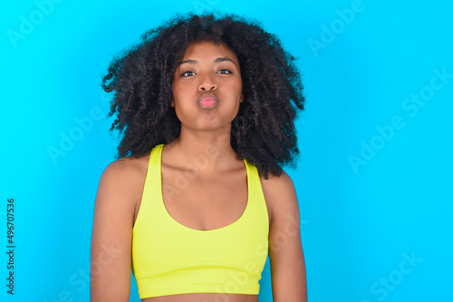 young woman with afro hairstyle in sportswear against blue background puffing cheeks with funny face. Mouth inflated with air, crazy expression. photo