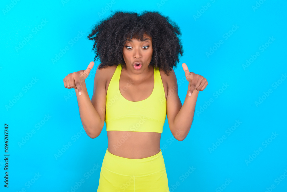 Amazed young woman with afro hairstyle in sportswear against blue background points down with fore fingers, opens mouth being shocked. Advertisement concept.