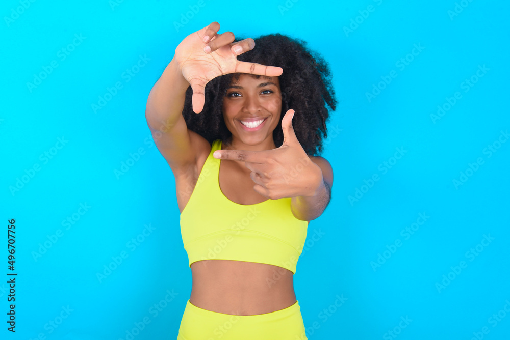 Positive young woman with afro hairstyle in sportswear against blue background with cheerful expression, has good mood, gestures finger frame actively at camera. Positive young woman with afro hairstyle in sportswear against blue background with cheerful expression, has good mood, gestures finger frame actively at camera.