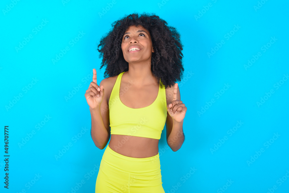 Successful friendly looking young woman with afro hairstyle in sportswear against blue background exclaiming excitedly, pointing both index fingers up, indicating something. Successful friendly looking young woman with afro hairstyle in sportswear against blue background exclaiming excitedly, pointing both index fingers up, indicating something.