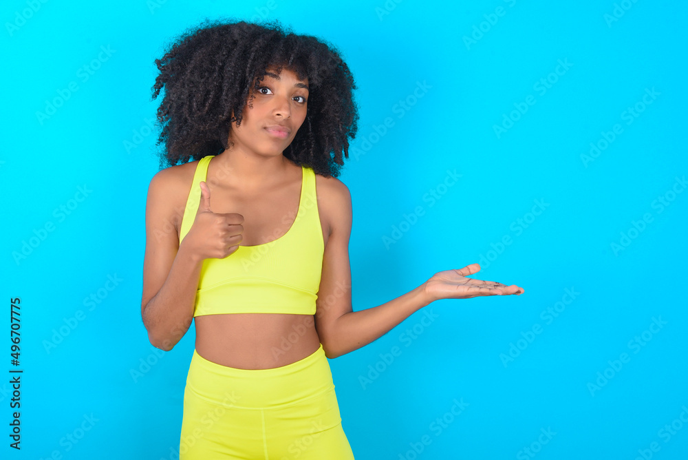 Happy cheerful young woman with afro hairstyle in sportswear against blue background showing thumb up and pointing with the other hand. I recommend this. Happy cheerful young woman with afro hairstyle in sportswear against blue background showing thumb up and pointing with the other hand. I recommend this.