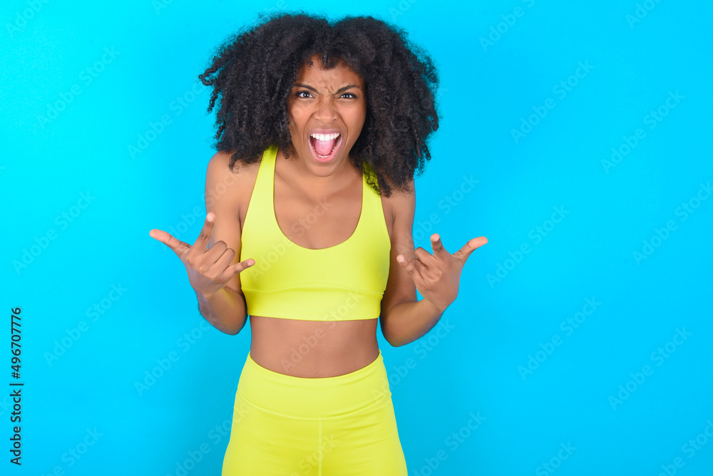 Born to rock this world. Joyful young woman with afro hairstyle in sportswear against blue background screaming out loud and showing with raised arms horns or rock gesture. Born to rock this world. Joyful young woman with afro hairstyle in sportswear against blue background screaming out loud and showing with raised arms horns or rock gesture.