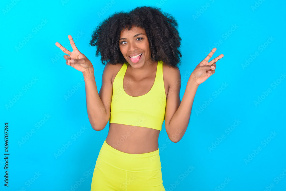 young woman with afro hairstyle in sportswear against blue background with optimistic smile, showing peace or victory gesture with both hands, looking friendly. V sign. young woman with afro hairstyle in sportswear against blue background with optimistic smile, showing peace or victory gesture with both hands, looking friendly. V sign.