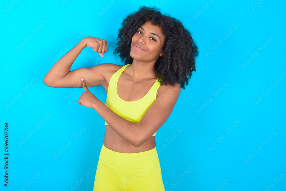 Smiling young woman with afro hairstyle in sportswear against blue background raises hand to show muscles, feels confident in victory, strong and independent. Smiling young woman with afro hairstyle in sportswear against blue background raises hand to show muscles, feels confident in victory, strong and independent.