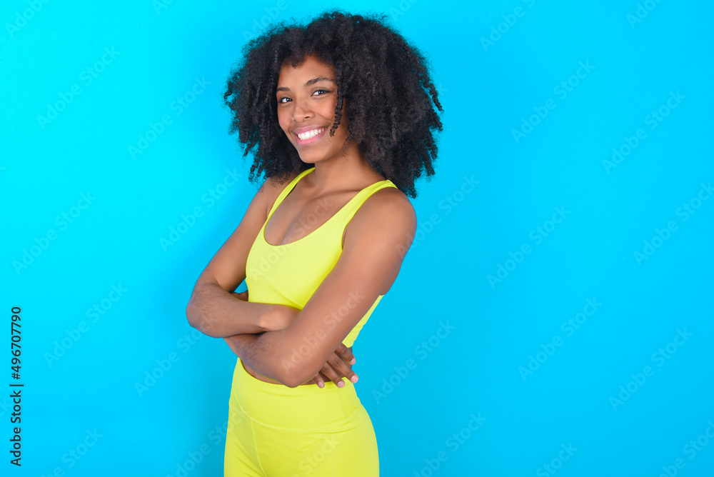 Image of cheerful young woman with afro hairstyle in sportswear against blue background with arms crossed. Looking and smiling at the camera. Confidence concept. Image of cheerful young woman with afro hairstyle in sportswear against blue background with arms crossed. Looking and smiling at the camera. Confidence concept.