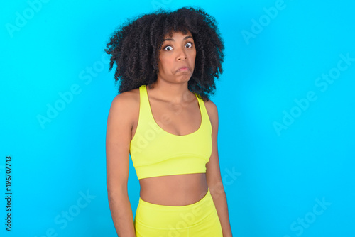 young woman with afro hairstyle in sportswear against blue background with snobbish expression curving lips and raising eyebrows, looking with doubtful and skeptical expression, suspect and doubt.