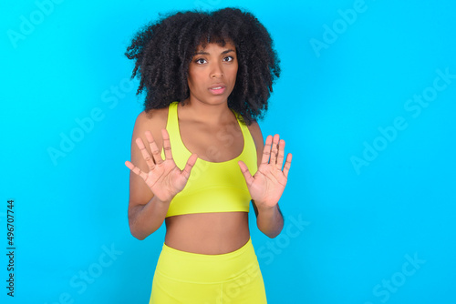 young woman with afro hairstyle in sportswear against blue background doing stop sing with palm of the hand. Warning expression with negative and serious gesture on the face.