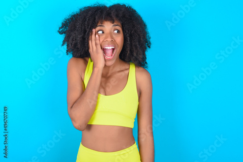 young woman with afro hairstyle in sportswear against blue background excited looking to the side hand on face. Advertisement and amazement concept.
