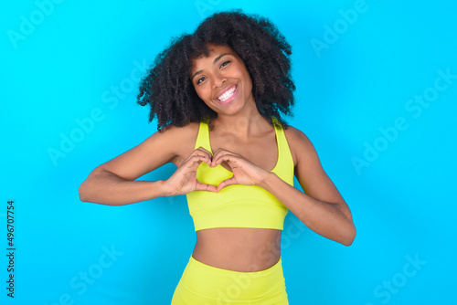 young woman with afro hairstyle in sportswear against blue background smiling in love showing heart symbol and shape with hands. Romantic concept.
