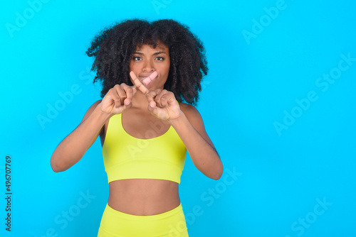 young woman with afro hairstyle in sportswear against blue background has rejection angry expression crossing fingers doing negative sign.