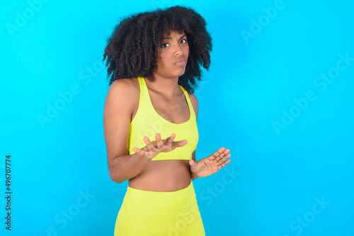 Afraid young woman with afro hairstyle in sportswear against blue background, makes terrified expression and stop gesture with both hands saying: Stay there. Panic concept.