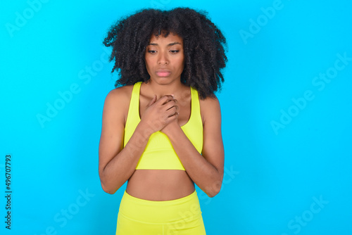 Sad young woman with afro hairstyle in sportswear against blue background feeling upset while spending time at home alone staring at camera with unhappy or regretful look.