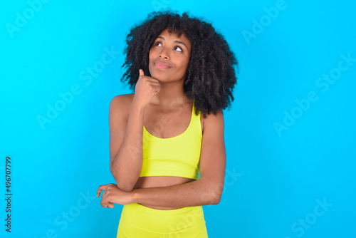 Face expressions and emotions. Thoughtful young woman with afro hairstyle in sportswear against blue background holding hand under his head, having doubtful look.